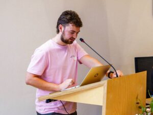 Thijmen staat op een spreekgestoelte voor een microfoon met laptop.